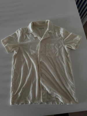 Sommarpiké från Kappahl - Helt oanvänd kortärmad pikéskjorta i krämvit färg från Kappahl. Skjortan har klassisk krage, knäppning framtill och en bröstficka. Tillverkad i ett mjukt, frottéliknande material som ger en avslappnad vibe. 