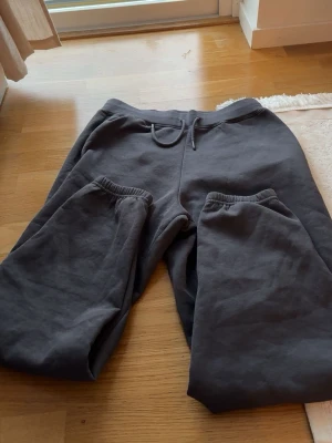 Mörkgrå mjukisbyxor med dragsko - Säljer ett par mörkgrå mjukisbyxor med resår i midjan och dragsko för skön passform. Byxorna har två sidofickor och muddar vid bensluten. Perfekta för chill dagar hemma eller när du vill vara bekväm.