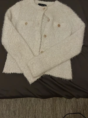 Vit fluffig kofta med guldknappar - Supersöt vit fluffig kofta med dekorativa guldknappar och två fejkade bröstfickor. Koftan har rund halsringning och är tillverkad i ett mjukt, lurvigt material som känns lyxigt. Perfekt att styla till jeans eller kjol för en trendig look.