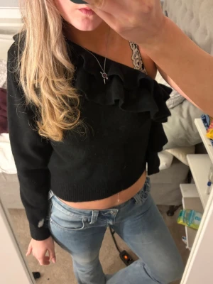 Svart croppad omlottopp med volang - Snygg svart stickad croppad topp med volangdetaljer och omlott-design över ena axeln. Toppen har lång ärm på ena sidan och är offshoulder på andra, vilket ger en trendig och asymmetrisk look. Perfekt för dig som vill sticka ut med din outfit. Helt ny❤️