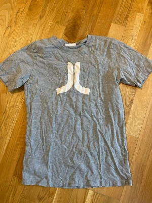 Grå Atari t-shirt med logga - Snygg grå t-shirt från Atari med den klassiska vita loggan tryckt på bröstet. T-shirten har rund hals och korta ärmar, perfekt för en avslappnad och cool stil. Tillverkad i mjuk bomull för skön komfort hela dagen.