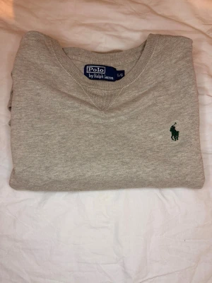 Beige sweatshirt från Polo Ralph Lauren - Klassisk beige tröja från Polo Ralph Lauren med rund halsringning och grönt broderat logotyp på bröstet. Tröjan har ribbade muddar och är tillverkad i mjukt material som passar perfekt till en avslappnad stil.