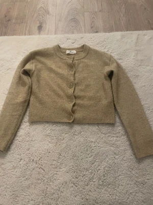 Beige kofta från H&M med knappar - Säljer en beige kofta från H&M med rund halsringning och stora matchande knappar framtill. Koftan är ribbstickad vid ärmslut och nederkant och har en croppad passform. Perfekt att slänga över en topp för en chill look.