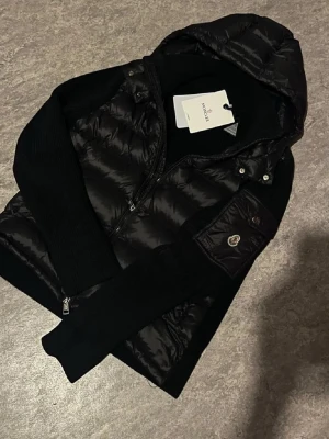 Svart pufferjacka från Moncler - Snygg svart pufferjacka från Moncler med glansig finish, huva och stickade ärmar. Jackan har dragkedja framtill och en ikonisk Moncler-logga på ärmen. Perfekt för kalla dagar när du vill hålla stilen. Var för liten när ja köpte den så inte använd! 