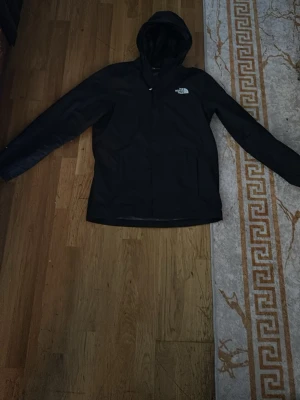Svart vindjacka från The North Face - Svart vindjacka från The North Face med huva och dragkedja framtill. Jackan har en stilren design med logga på bröstet och är tillverkad i ett lätt och tåligt material som skyddar mot vind.