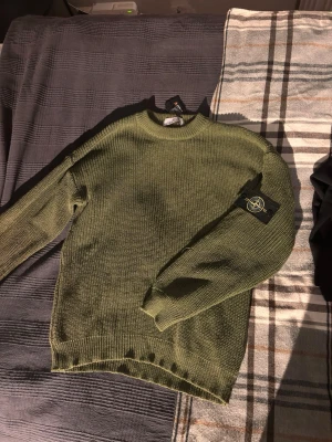 Olivgrön stickad tröja Stone Island - Säljer en olivgrön stickad tröja från Stone Island med klassisk patch på vänster ärm. Tröjan har rund hals, ribbade muddar och är gjord i grovstickad bomull. Perfekt för dig som gillar streetwear och vill ha något stilrent och bekvämt.