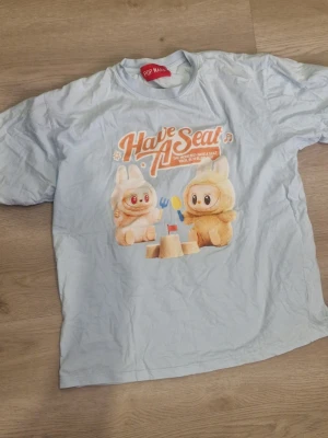 Ljusblå Pop Mart T-shirt med tryck - Ljusblå T-shirt från Pop Mart med ett färgglatt tryck av två gulliga labbobo texten 'Have A Seat'. T-shirten har rund hals och korta ärmar. Aldrig använt vilket gillar inte de länger. Står ingen storlek men mellan (cirka 155 till 163)