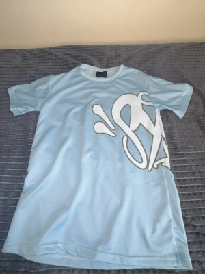 Ljusblå Syna Wolrd t-shirt med stor vit print - Säljer en ljusblå t-shirt med stor vit grafisk print både fram och bak. T-shirten har korta ärmar och en klassisk rund halsringning. Materialet känns mjukt och skönt, troligtvis bomull eller en bomullsblandning. Perfekt för dig som gillar streetwear vibes.