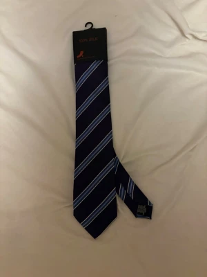 Mörkblå sidenslips med ränder - Snygg mörkblå slips i 100% siden med diagonala ränder i ljusblått och vitt. Klassisk design som passar till skjorta och kavaj. Perfekt accessoar för att lyfta din outfit med en elegant touch.