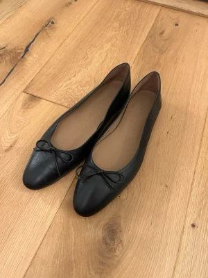 Svarta ballerinaskor med rosett - Stilrena svarta ballerinaskor i mjukt läder med klassisk rund tå och liten rosett framtill. Använda en gång men säljer då de inte passar!