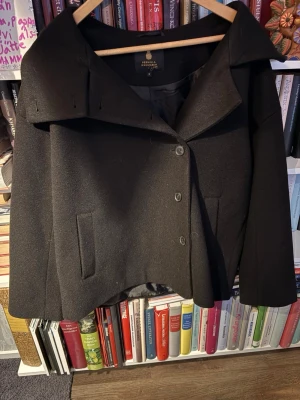 Svart ullkappa Pernilla Wahlgren - Stilren svart kappa från Pernilla Wahlgren med bred krage och tre knappar framtill. Kappan har raka fickor och är tillverkad i ett tjockare ullmaterial som håller värmen. Kortare modell som ger en modern och trendig look.