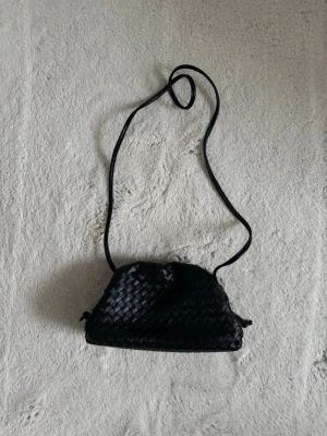 Svart flätad axelväska i skinnimitation - Snygg svart axelväska med flätad design och laxelrem. Väskan har en mjuk, lite trekantig form och är gjord i skinnimitation. Perfekt för dig som gillar stilrena accessoarer med en twist. Den är från jacki, använd fåtal gånger och ordinarie pris är 2900kr