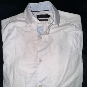 Vit skjorta från Riley - Klassisk vit skjorta från Riley med lång ärm och knappar framtill. Skjortan har en snygg kontrastdetalj i ljusblått mönster på insidan av kragen och knappslån. Perfekt för en clean och stilren look. Skjortan är ”super slim fit” storlek xs 