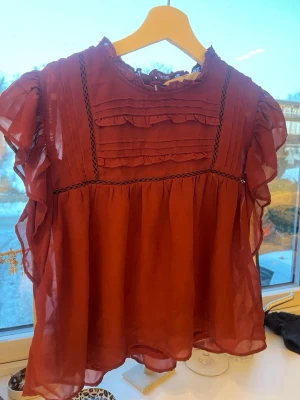 Vinröd volangblus med spetsdetaljer - Söt vinröd blus i transparent material med volanger på ärmarna och spetsdetaljer framtill. Blusen har rund halsringning, knäppning bak i nacken och dekorativa veck på bröstet. Perfekt för dig som gillar romantisk och trendig stil. Från hm storlek 158 (12-14 år)