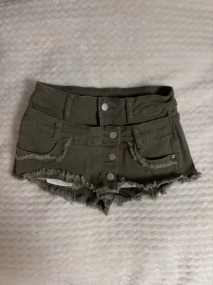 Olivgröna shorts med knappar - Säljer ett par olivgröna shorts med fransig kant och fyra silverfärgade knappar framtill. Shortsen har fickor både fram och bak samt råa detaljer längs kanterna för en avslappnad look. Perfekta för sommaren!