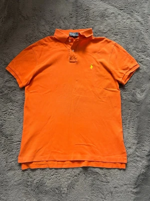 Orange pikétröja från Polo Ralph Lauren - Klassisk orange pikétröja från Polo Ralph Lauren med korta ärmar och krage. Tröjan har två knappar vid halsen och den ikoniska gula logotypen broderad på bröstet. Tillverkad i mjuk bomull för en bekväm känsla.