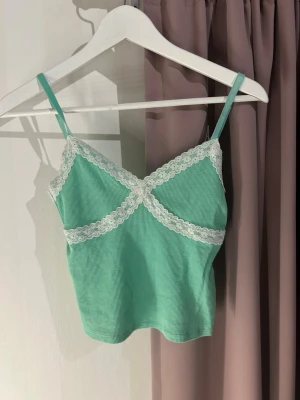 Mintgrönt ribbat linne med spets - Säljer ett mintgrönt ribbat linne med smala axelband och vit spetsdetalj längs urringningen och under bysten. Toppen har en croppad passform och är perfekt för varma dagar eller som layering-piece. Supersöt och trendig stil!