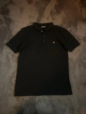 Svart pikétröja från Lyle & Scott - Klassisk svart pikétröja från Lyle & Scott med korta ärmar och gul broderad logga på bröstet. Tröjan har krage och knäppning med tre knappar framtill. Perfekt till jeans eller chinos för en clean och stilren look.