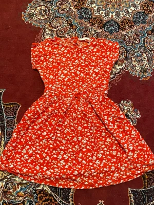 Röd blommig klänning med kort ärm - Söt röd klänning med små vita blommor och korta ärmar. Klänningen har rund halsringning och markerad midja med utställd kjol. Perfekt för dig som gillar färgglada och somriga plagg. Materialet känns lätt och luftigt.