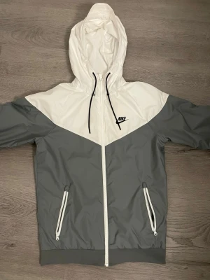 Nike gråvit vindjacka med huva - Nike vindjacka i grått och vitt med dragkedja framtill, två fickor med vita dragkedjor och justerbar huva med svarta snören. Jackan har en sportig look med logga på bröstet och elastiska muddar vid ärmar och midja. Perfekt för blåsiga dagar.
