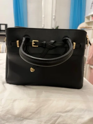 Svart handväska med gulddetaljer - Säljer en handväska som inte kommer till användning längre men har några fläckar på insidan. Vet dock inte vart den är ifrån.