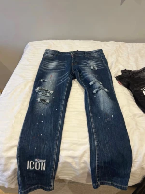 Dsquared2 ICON slitna jeans blå - Säljer ett par blå Dsquared2 ICON jeans med slitningar, hål och färgstänk. Jeansen har en rak passform, klassisk femficksdesign och tydlig logga vid benslutet. Perfekta för dig som gillar en edgy och trendig streetstil.