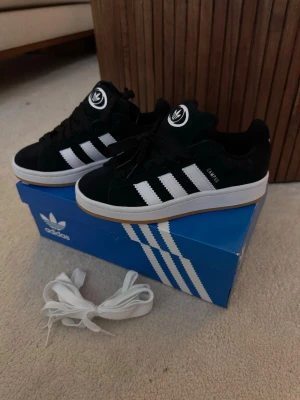Adidas Campus svarta sneakers - Svarta Adidas Campus sneakers i mocka med klassiska vita ränder på sidorna och vit sula med gummidetalj. Tjock plös med Adidas-logga och extra vita skosnören medföljer. Snygg retrostil som funkar till många outfits.