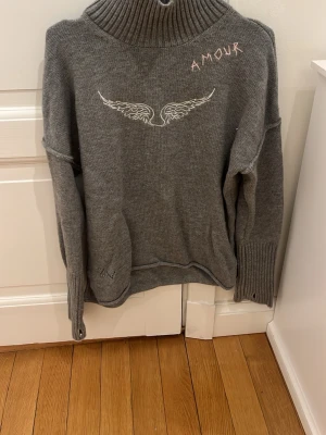 Grå stickad tröja  - Mysig grå stickad tröja med hög krage och broderad vinge framtill samt texten 'AMOUR' i rosa. Tröjan har långa ärmar och ribbstickade muddar. Perfekt för kalla dagar och enkel att matcha med jeans eller kjol.har användt den typ 5 gånger och köpte den för nästan 2000kr❤️