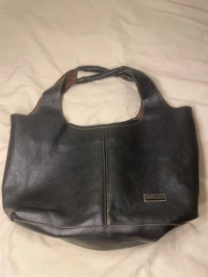 Svart läderhandväska från Jimmy Choo - Stilren svart handväska i äkta läder från Jimmy Choo med bruna detaljer på insidan. Väskan har dubbla handtag, dekorativ söm framtill och en metallogo. Invändigt finns flera fack och dragkedja för smart förvaring.