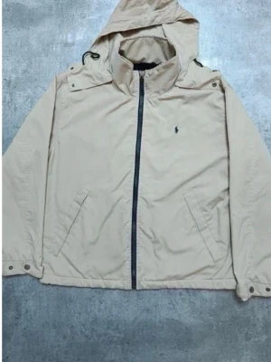 Beige vindjacka från Polo Ralph Lauren - Stilren beige vindjacka från Polo Ralph Lauren med klassisk marinblå logga på bröstet. Jackan har huva, dragkedja framtill, snedställda fickor och justerbara muddar. Perfekt för blåsiga dagar och enkel att matcha med olika outfits.