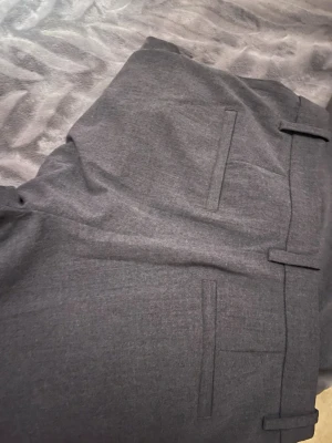 Grå kostymbyxor - Snygga grå kostymbyxor med klassisk rak passform och bälteshällor. Byxorna har två bakfickor med lock och är tillverkade i ett mjukt tyg som ger en stilren look. Perfekta för dig som vill ha en clean och tidlös stil.