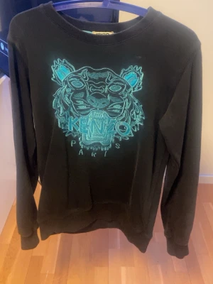 Svart Kenzo sweatshirt med tiger - Säljer en svart sweatshirt från Kenzo med ikonisk tigerbrodyr i turkost och blått framtill. Tröjan har rund halsringning, långa ärmar och ribbade muddar. Perfekt för dig som vill ha en statement piece i garderoben.