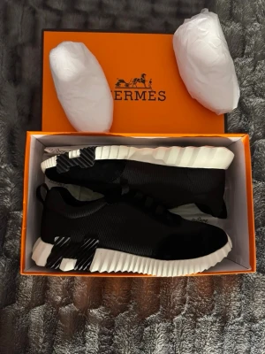 Svarta H sneakers med vit sula - Snygga svarta sneakers från H med en chunky, räfflad vit sula och svarta detaljer på hälen. Skorna har en modern design med stickad ovandel och diskret branding. Perfekta för dig som gillar exklusiva och trendiga sneakers.