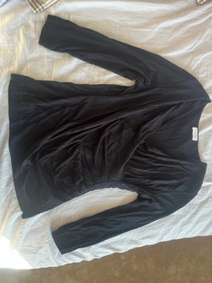 Svart långärmad topp omlott - En svart långärmad topp med omlott-skärning och smal passform. Toppen har en enkel, stilren design och är tillverkad i ett mjukt material som känns skönt mot huden. Perfekt att styla med jeans eller kjol för en clean look.