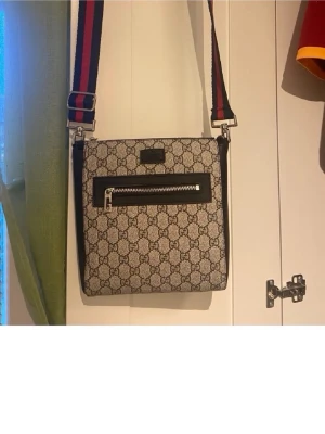Gucci axelväska med monogram och bred rem - Snygg axelväska från Gucci i beige canvas med klassiskt GG-monogram och svarta läderdetaljer. Väskan har en bred axelrem i rött och marinblått, samt en praktisk ytterficka med dragkedja framtill. Perfekt för dig som gillar streetstyle och designeraccessoarer.  Mycket bra skick. Priset kan diskuteras 