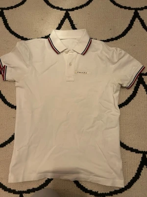 Vit piké från Prada med röd detalj - Vit pikétröja från Prada med klassisk krage och knäppning framtill. Röd och marinblå rand på krage och ärmslut ger en sportig touch. Diskret Prada-logga på bröstet. Perfekt för en stilren och trendig look.