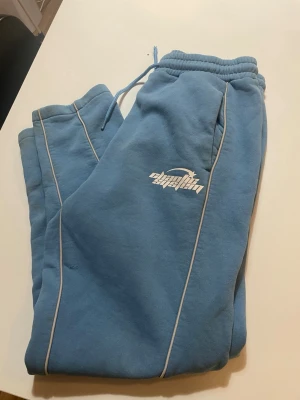 Blå mjukisbyxor från Planet System - Säljer ett par ljusblå mjukisbyxor från Planet System med vita kontrastsömmar och logga på benet. Byxorna har dragsko i midjan och muddar vid bensluten. Perfekta för chill dagar eller när du vill vara extra bekväm.