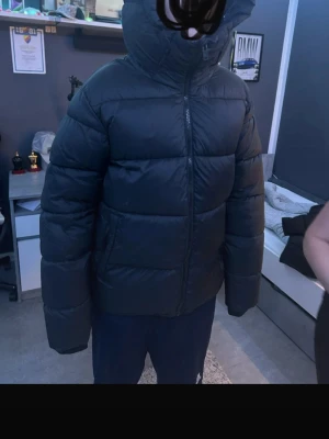 Svart pufferjacka med huva - Säljer en svart pufferjacka med huva och dragkedja framtill. Jackan har en quiltad design och ett diskret märke på bröstet. Perfekt för kalla dagar och riktigt varm tack vare den tjocka vadderingen.