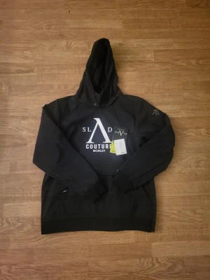 Svart skidjacka från Slade Couture - Snygg svart skidjacka från Slade Couture med stor logga och text på bröstet samt patch på ärmen. Tröjan har huva och ribbade muddar vid ärmslut och nederkant. Perfekt för sportlovet och riktigt bekväm att bära.