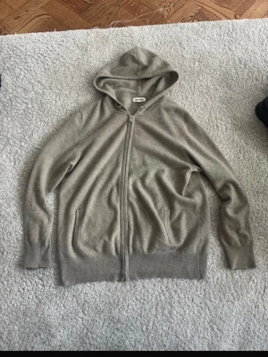 Grå hoodie i kashmir från Soft Goat - Mysig grå hoodie från Soft Goat i mjuk kashmir. Dragkedja framtill, två fickor och ribbade muddar vid ärmslut och nederkant. Perfekt för chill dagar och enkel att matcha med allt.