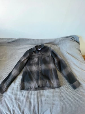 Grå overshirt från Jack & Jones - Snygg grå overshirt från Jack & Jones med diskret rutmönster. Skjortan har två bröstfickor med lock, knäppning framtill och klassisk krage. Perfekt lager på lager-plagg för en avslappnad stil. Tillverkad i mjukt tyg som känns bekvämt hela dagen.