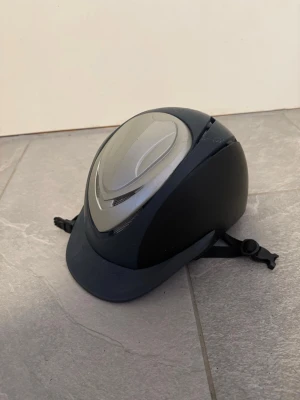 Svart och grå ridhjälm - Snygg ridhjälm i svart och grått med justerbart hakband och ventilerande detaljer på ovansidan. Hjälmen har en klassisk form med skärm framtill och är tillverkad i tålig plast med mesh för ventilation.