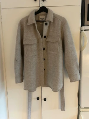 Gråbeige kappa - Gråbeige kappa med bälte. Har en svag fläck på ena armen, men skulle säga att den syns bättre på bild än i verkligheten.