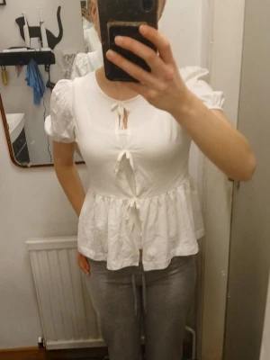 Vit topp med puffärm och knyt - Söt vit topp med puffärmar och tre knyt framtill. Modellen har en lätt utsvängd peplum nedtill och rund halsringning. Perfekt för en trendig och feminin look. Tunt och luftigt material, passar bra till jeans eller kjol.