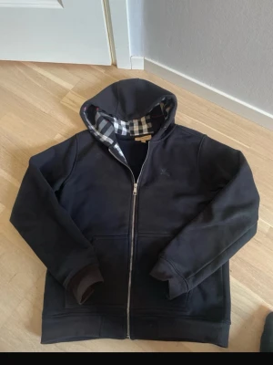 Svart kofta från Burberry med dragkedja - Svart kofta från Burberry med dragkedja framtill och klassiskt rutigt foder i huvan. Hoodien har två fickor fram och ribbade muddar vid ärmar och nederkant. Perfekt för dig som gillar stilrena plagg med exklusiv känsla och snygga detaljer. Size M men sitter mer som S