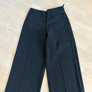 Svarta vida kostymbyxor - Snygga svarta kostymbyxor med hög midja och vida ben. Byxorna har pressveck framtill och stängs med dragkedja och hake. Perfekta för en stilren och trendig look. Materialet känns slätt och klassiskt.