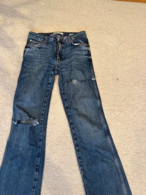 Blå slitna jeans från Zara - Säljer ett par blå jeans från Zara med slitningar och hål vid knäna för en trendig och avslappnad look. Jeansen har klassisk femficksmodell, normal midja och raka ben. Perfekta för dig som gillar en edgy stil.