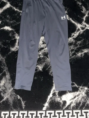 Grå träningsbyxor från Under Armour - Säljer ett par grå träningsbyxor från Under Armour med resår och dragsko i midjan. Byxorna har en enkel och stilren design med logga på låret och är tillverkade i ett lätt och snabbtorkande material, perfekt för gym eller löpning.