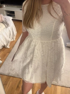 Vit spetsklänning med v-ringning - Söt vit kortklänning i spets med v-ringning i ryggen och halvlånga ärmar. Klänningen har en figurnära överdel och utställd kjol, perfekt för sommarens alla tillfällen speciellt studsanden!
