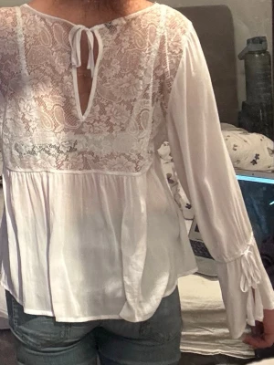 Vit blus med spets och knytband - Säljer en luftig vit blus med vackra spetsdetaljer upptill och knytband vid halsen. Blusen har långa, vida ärmar med dekorativa band vid ärmsluten och en lätt v-ringning. Den är lite stor på mig och jag har vanligtvis xs i tröjor. Så jag gissar på storleken S-M. Perfekt för en romantisk och bohemisk stil.